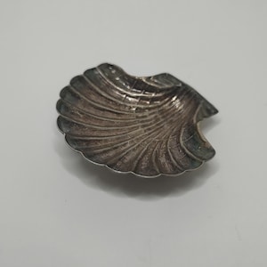 May include: A small, antique silver-colored seashell-shaped dish. The dish has a detailed, ridged texture and a scalloped edge. The dish is likely made of metal and is displayed on a white surface.