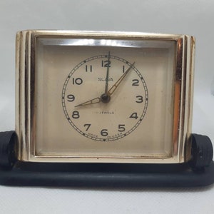 May include: A silver-colored alarm clock with a white face and black hands. The clock is labeled "SLAVA" and "II JEWELS". The clock is made in the USSR.