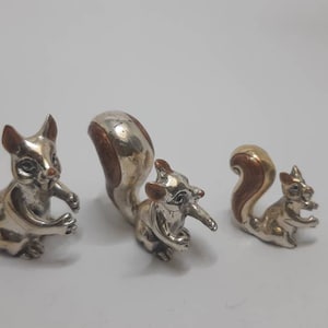 May include: Three silver and gold squirrel figurines of varying sizes. The largest squirrel is silver with a brown tail, the middle squirrel is silver with a brown tail, and the smallest squirrel is gold with a brown tail.