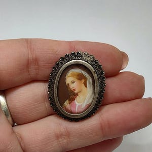 May include: A silver oval brooch with a detailed filigree design. The brooch features a painted portrait of a woman with a white head covering. The woman is wearing a pink dress and has a light brown hair.