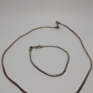 Puede incluir: Un juego de collar y pulsera de cadena de serpiente plateada. El collar es más largo que la pulsera y ambos tienen un acabado brillante.