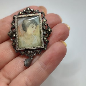 May include: A silver and marcasite brooch with a cameo of a woman in a white dress. The cameo is set in a square frame with a decorative border. The brooch has a dangling marcasite drop.