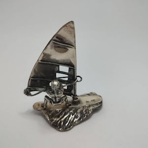 Puede incluir: Una figurita de plata de una persona haciendo windsurf en una ola. La figurita es detallada y tiene un aspecto realista.