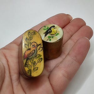 Puede incluir: Dos pequeñas cajas de baratijas ovaladas de latón con diseños de pájaros pintados. Una caja presenta un pájaro amarillo con marcas negras sobre un fondo blanco, la otra caja presenta un pájaro rojo y naranja sobre un fondo amarillo.