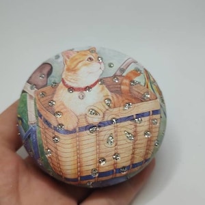 Puede incluir: Un adorno redondo de cerámica con una imagen de un gato sentado en una cesta de mimbre. El adorno está decorado con diamantes de imitación.