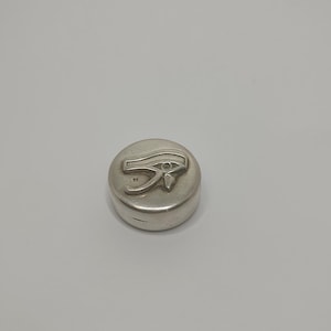 May include: A silver-colored, round container with a raised Eye of Horus symbol on the top. The container is made of a shiny, metallic material and sits on a plain, light gray surface. The design is a classic Egyptian motif.