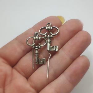May include: Two silver-colored key charms with intricate details, held in a hand. The keys have ornate handles and rectangular key teeth. The background is a plain white surface. These charms could be used for jewelry or craft projects.