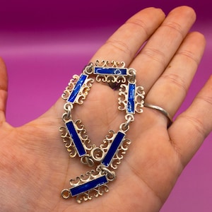 May include: Silver-toned bracelet with rectangular blue enamel inlays. The bracelet features ornate, openwork designs between the enamel sections. The clasp is a simple hook and eye closure. The bracelet is displayed against a pink background.