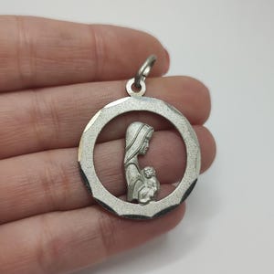 May include: Silver pendant featuring a depiction of the Virgin Mary holding the baby Jesus within a circular frame. The pendant has a textured border and a small loop for a chain.