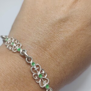 May include: A silver bracelet with a floral design and green accents. The bracelet features a series of interconnected floral-shaped links, each with a small green stone in the center. The bracelet is shown on a person's wrist.