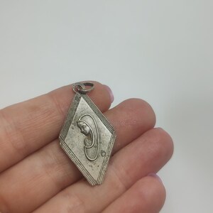May include: A silver pendant in the shape of a diamond with a raised image of the Virgin Mary praying inside. The pendant has a loop at the top for attaching to a chain.