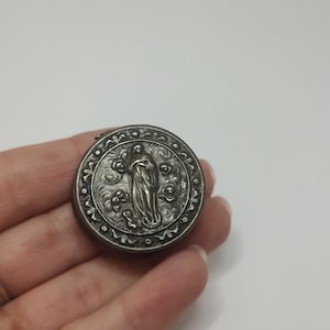 Puede incluir: Un colgante religioso redondo y plateado antiguo. El colgante presenta un relieve detallado de una figura, posiblemente un santo o un icono religioso, rodeado de elementos florales y decorativos. El diseño es intrincado y detallado.