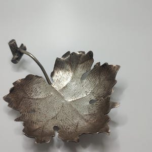 Könnte beinhalten: Eine silberfarbene, blattförmige Schale mit strukturierter Oberfläche. Die Schale hat einen Stiel und ein kleines Astdetail. Das Blatt hat ein detailliertes Adernmuster und ist wahrscheinlich aus Metall.