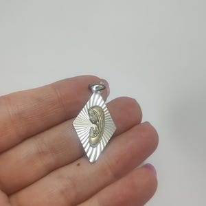 May include: Silver-tone pendant with a gold-colored religious figure in the center. The pendant is diamond-shaped with a sunburst design. The figure is a depiction of a religious icon.