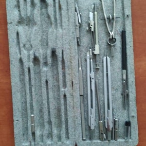 May include: A set of drafting tools in a gray case. The tools include compasses, rulers, and a protractor. The tools are made of metal and have a silver finish.