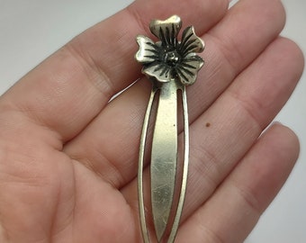May include: Silver bookmark with a flower design. The flower is in the center of the bookmark and has five petals. The bookmark is about 5 inches long.