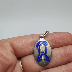 May include: A silver oval pendant with a blue enamel image of a woman in prayer. The woman is wearing a blue robe and has a white face.