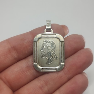 May include: A silver pendant with a rectangular frame and a detailed profile of a person with long hair and a beard. The pendant has a small loop at the top for a chain.