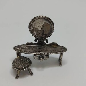 May include: A silver vanity set with a round mirror, a small table with a drawer, and a chair. The set is made of silver and has a vintage look.