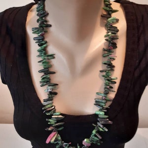 Op de afbeelding: Een lange ketting gemaakt van groene en roze edelsteenchips. De ketting is gemaakt van veel kleine, onregelmatig gevormde chips die aan elkaar geregen zijn op een dun koord. De ketting is ongeveer 50 cm lang.