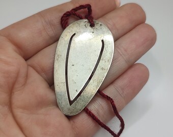 May include: Silver-toned metal bookmark with a cut-out design and a dark red cord. The bookmark has a smooth, polished surface and a teardrop shape. A simple and elegant accessory for book lovers.