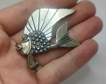 May include: A silver-toned fish brooch with a textured body and a detailed fin design. The fish has a raised eye and a slightly open mouth.