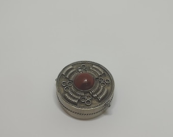May include: A small, round, silver-colored box with a central, reddish-brown stone. The box has an intricate, circular design with a textured surface and a decorative border. The lid is adorned with a central stone and surrounding circular patterns.
