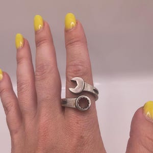 Puede incluir: Un anillo de metal en tono plateado con forma de llave inglesa en miniatura que se envuelve alrededor del dedo. El diseño presenta una cabeza de llave abierta en un lado y una cabeza de llave cerrada en el otro, creando una banda continua.