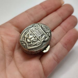 May include: A silver oval-shaped trinket box with an intricate design featuring a beetle and floral motifs.