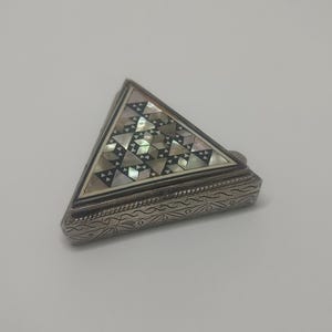 Op de afbeelding: Een zilverkleurige, driehoekige doos met een scharnierend deksel. Het deksel is ingelegd met een geometrisch patroon van iriserende parelmoer driehoeken. De basis van de doos heeft een gegraveerd, herhalend golfpatroon. Een kleine, ronde knop zit aan de zijkant.