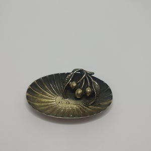 May include: A small, silver-colored dish with a decorative design of three gold-colored cherries on a branch. The dish has a scalloped edge and a textured surface.