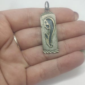 May include: A rectangular silver pendant with a stylized floral design in blue and gold. The pendant has a small loop at the top for a chain. The edges of the pendant have a textured border.