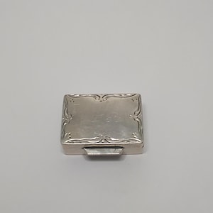 Può includere: Una piccola scatola rettangolare d'argento con volute decorative sui bordi. La scatola ha un coperchio incernierato e una piccola chiusura rettangolare. L'argento ha una finitura lucida, che riflette la luce. La scatola sembra essere vintage o antica.