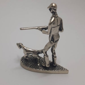 May include: Silver figurine of a hunter with a gun and a dog. The hunter is wearing a hat and the dog is running beside him. The figurine is on a heart-shaped base.
