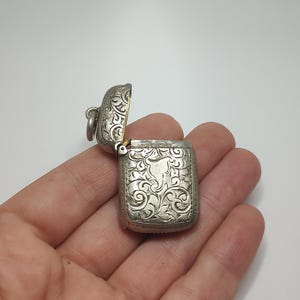 May include: A small, silver-colored, rectangular box with an open lid. The box and lid are intricately engraved with a swirling floral pattern. The box is held open, revealing the interior. The box has a small loop on the side.