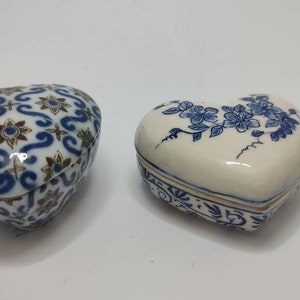 Op de afbeelding: Twee hartvormige keramische sieradendoosjes met blauwe en witte bloemenmotieven. Een doosje heeft een ingewikkelder patroon met gouden accenten.
