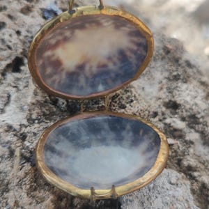 Może przedstawiać: Otwarta zawieszka w kształcie muszli ze złotą ramką. Wnętrze zawieszki ma naturalny wzór w odcieniach brązu, bieli i błękitu. Mała pętla jest przymocowana u góry do zawieszenia. Zawieszka jest na zawiasach.