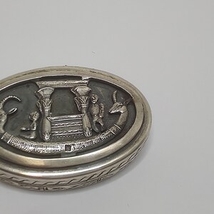 May include: A silver oval trinket box with an intricate design featuring an Egyptian-style temple with two figures and a gazelle. The box has a detailed border.