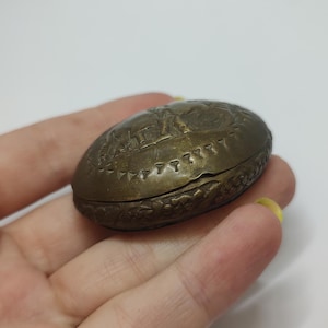May include: A small, round, brass trinket box with an intricate design. The box is closed and features a raised design of a scene with figures and a border of small triangles.