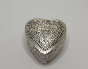 May include: A silver heart-shaped trinket box with intricate floral and scrollwork designs. The box is closed and sits on a white surface. The lid and sides are decorated with detailed patterns.