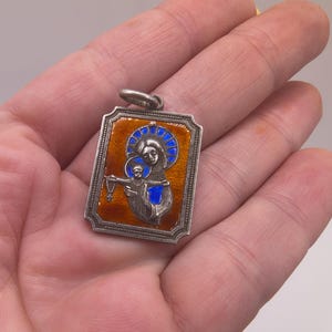 May include: A rectangular silver pendant featuring a raised relief of a figure holding a child, accented with vibrant orange and blue enamel. The metal frame has a decorative border and a top loop for attachment, showcasing intricate religious imagery.