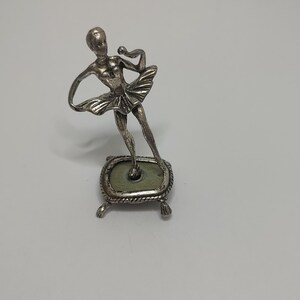 Puede incluir: Una figurita de plata de una bailarina de pie sobre una pequeña base acolchada verde. La bailarina lleva una falda y tiene los brazos levantados.