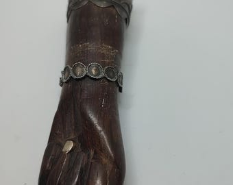 May include: A wooden hand sculpture with silver accents. The hand is carved in a closed fist position with a silver band around the wrist and a silver band around the top of the hand. The hand is dark brown in color.