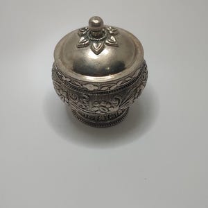 May include: A small, silver-colored decorative container with a lid. The container has intricate floral and geometric patterns etched into its surface. The lid is topped with a small, round knob. The container is likely made of metal.