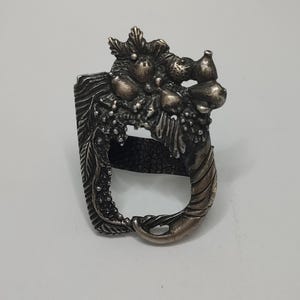 May include: A silver napkin ring with a detailed design of a fruit-filled cornucopia. The ring is made of metal and has a textured surface.