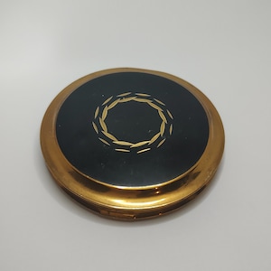 Puede incluir: Un espejo compacto con borde dorado, superficie negra y diseño de corona dorada.