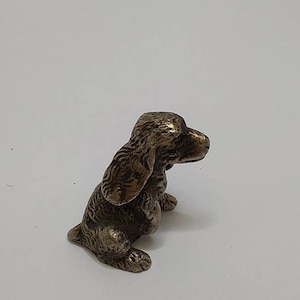 May include: A small, bronze figurine of a puppy sitting with its head tilted to the side. The puppy has a detailed, textured coat and a sweet expression.