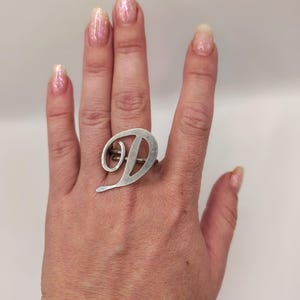 Op de afbeelding: Een zilverkleurige ring met een grote, gestileerde hoofdletter D in een cursief lettertype. De letter is horizontaal over de vinger geplaatst, waarbij de metalen band achter het letterontwerp zichtbaar is, wat zorgt voor een opvallend sieraad.