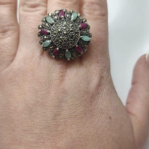 Può includere: Un anello d'argento con un designo rotondo a forma di fiore. L'anello è incastonato con piccole pietre grigio scuro e accentato con pietre rosa e verdi.