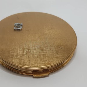 Puede incluir: Un espejo compacto dorado con una letra "S" plateada en la tapa.